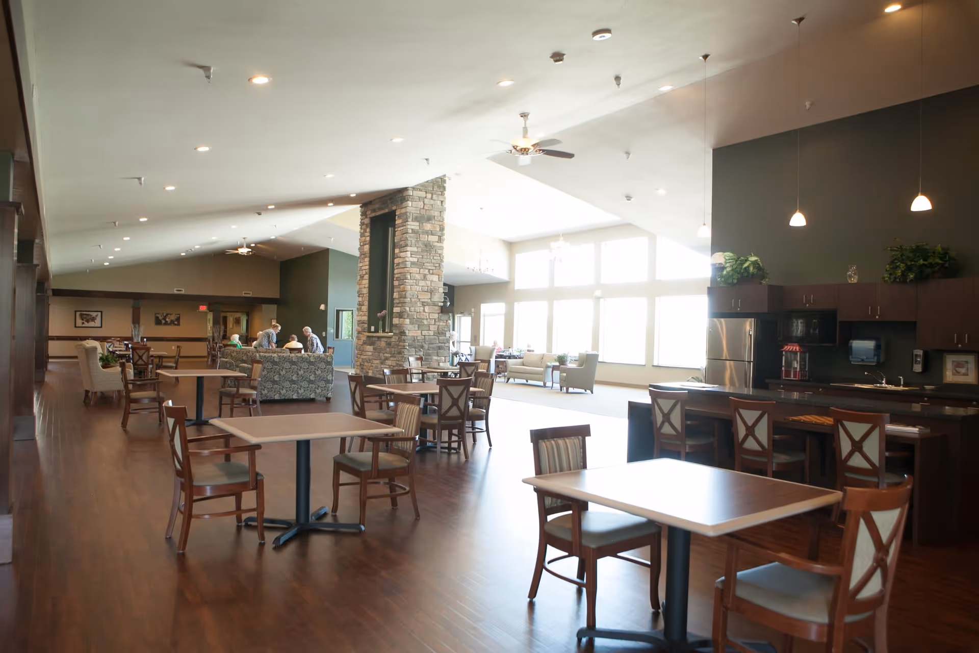Spacious and well-lit common area in an assisted living facility featuring multiple wooden tables and chairs, a stone fireplace in the center, a kitchen area with a refrigerator and cabinets on the right, and several elderly people seated and conversing in the background near large windows.