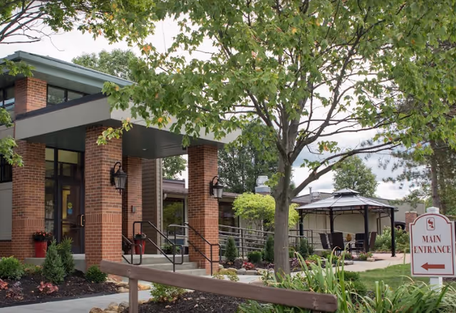 Exterior view of Cardinal Retirement Village showing the main entrance with brick pillars, a covered walkway, a tree in the foreground, landscaped garden beds, and a gazebo with seating in the background.