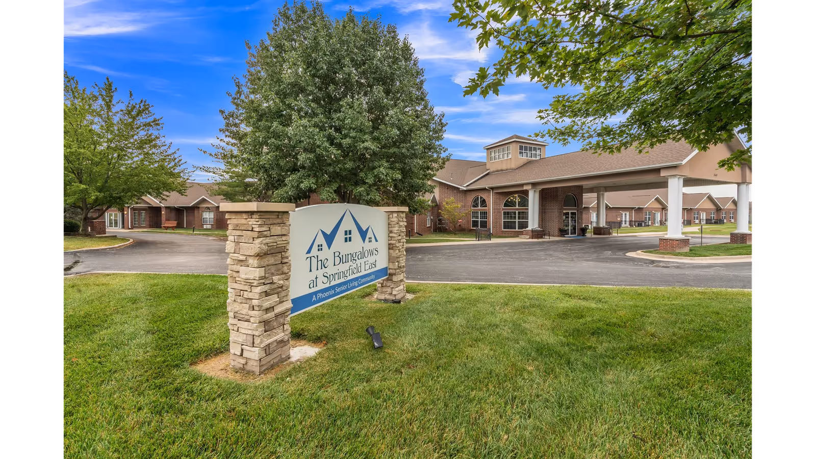 Exterior view of The Bungalows at Springfield East senior living community showing a stone and signboard entrance with the facility name, surrounded by green grass and trees, with the building and covered driveway visible in the background under a partly cloudy blue sky.
