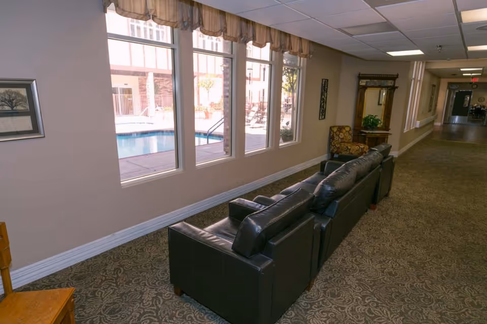 Interior hallway of an assisted living facility with a black leather couch and an armchair near a window. The window shows a view of an outdoor swimming pool area. The hallway has patterned carpet, beige walls, and ceiling lights.