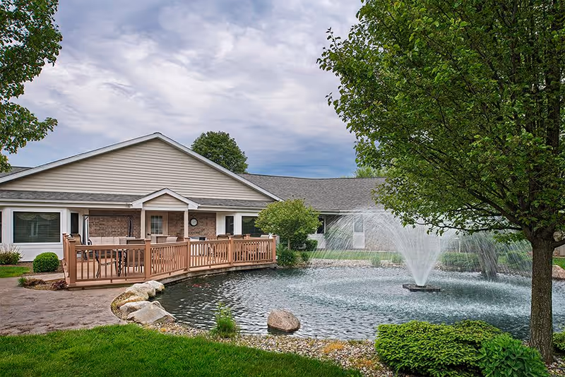 A peaceful outdoor scene at American House Holland featuring a pond with a large water fountain in the center, surrounded by green grass, bushes, and trees. A building with a beige exterior and a wooden deck with patio furniture is visible in the background under a cloudy sky.