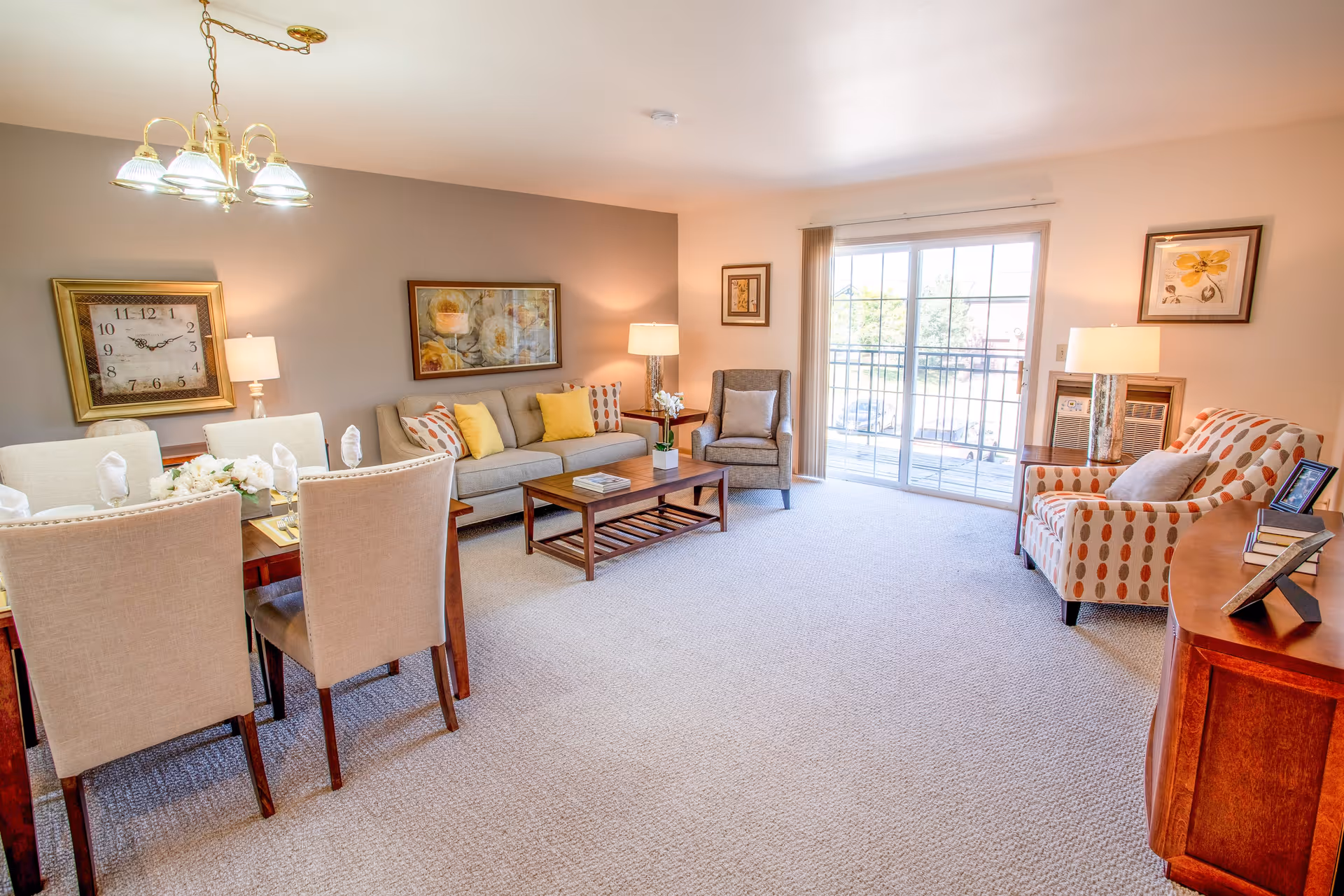 A bright and spacious living room and dining area in a senior living facility. The room features a beige carpet, a wooden dining table with six upholstered chairs, a beige sofa with yellow and patterned cushions, a wooden coffee table, two armchairs, and several framed artworks on the walls. Large sliding glass doors lead to an outdoor balcony, allowing natural light to fill the space.