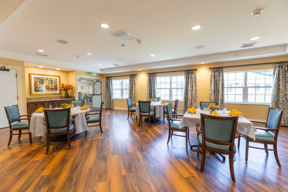 A bright dining room with wooden floors, several round tables covered with white tablecloths, each set with yellow napkins, glassware, and floral centerpieces. The room has large windows with patterned curtains allowing natural light to fill the space. There is a painting and a vase with flowers on a sideboard against one wall.
