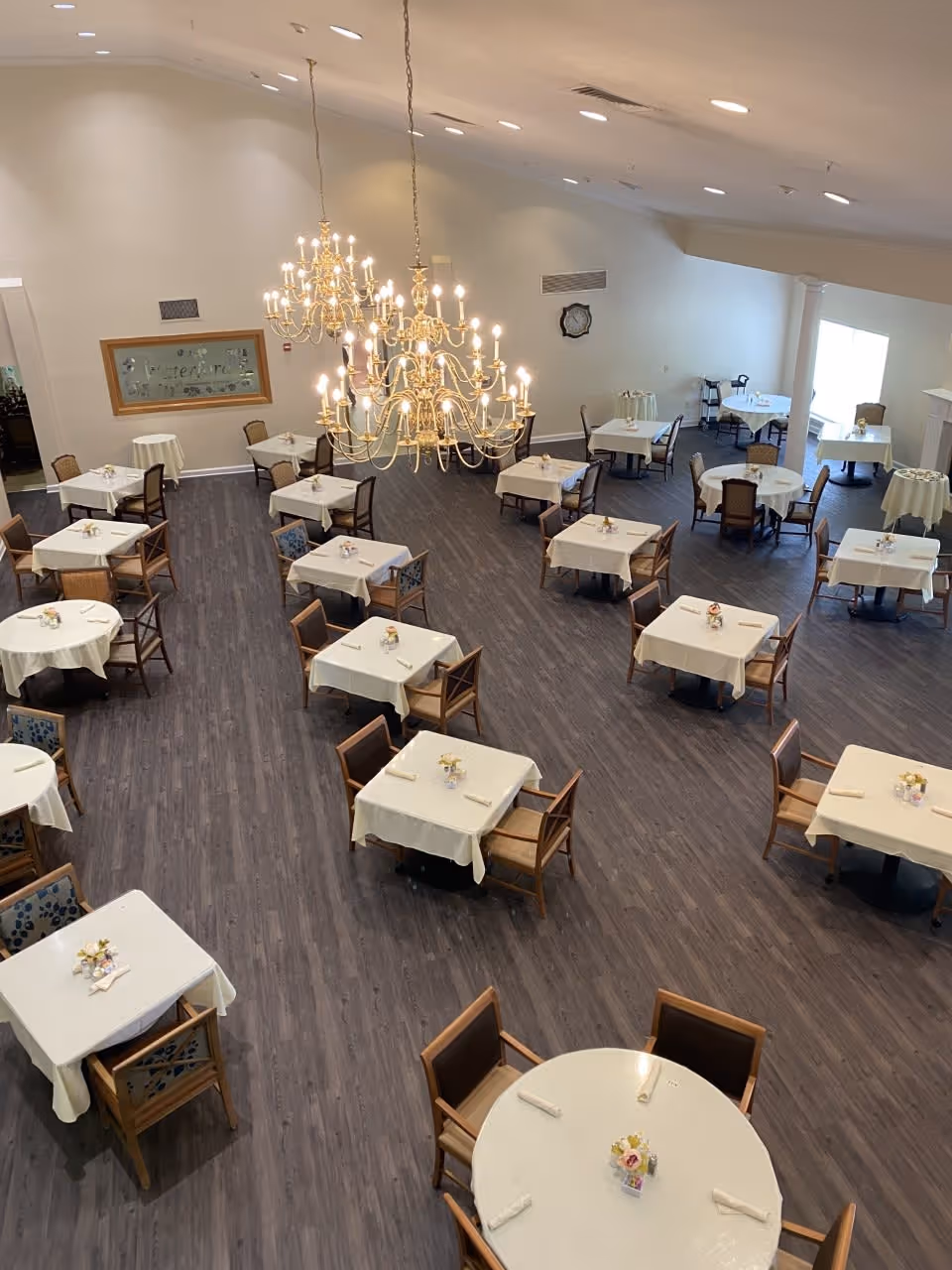 A spacious dining room with multiple tables covered in white tablecloths, each set with napkins and small floral centerpieces. The room features dark wood flooring, wooden chairs, and two large ornate chandeliers hanging from the ceiling. There is a framed sign on the wall and a clock, with natural light coming in from windows on the right side.