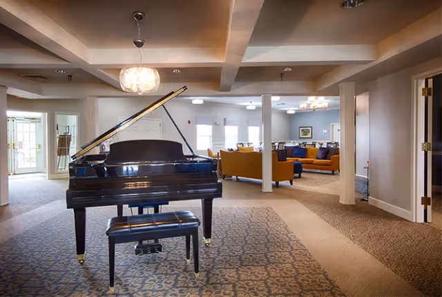 A spacious communal lounge with a black grand piano in the foreground and seating areas with sofas in the background.