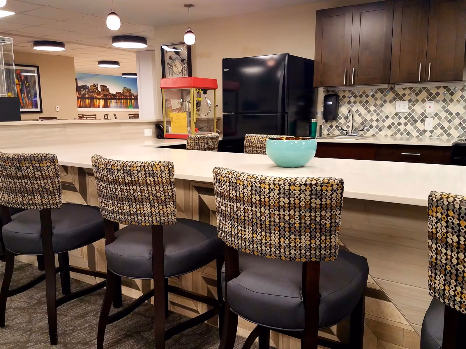 Communal kitchen and bar area with patterned barstools at a white countertop, a teal bowl, black refrigerator, cabinets and a popcorn machine in the background.