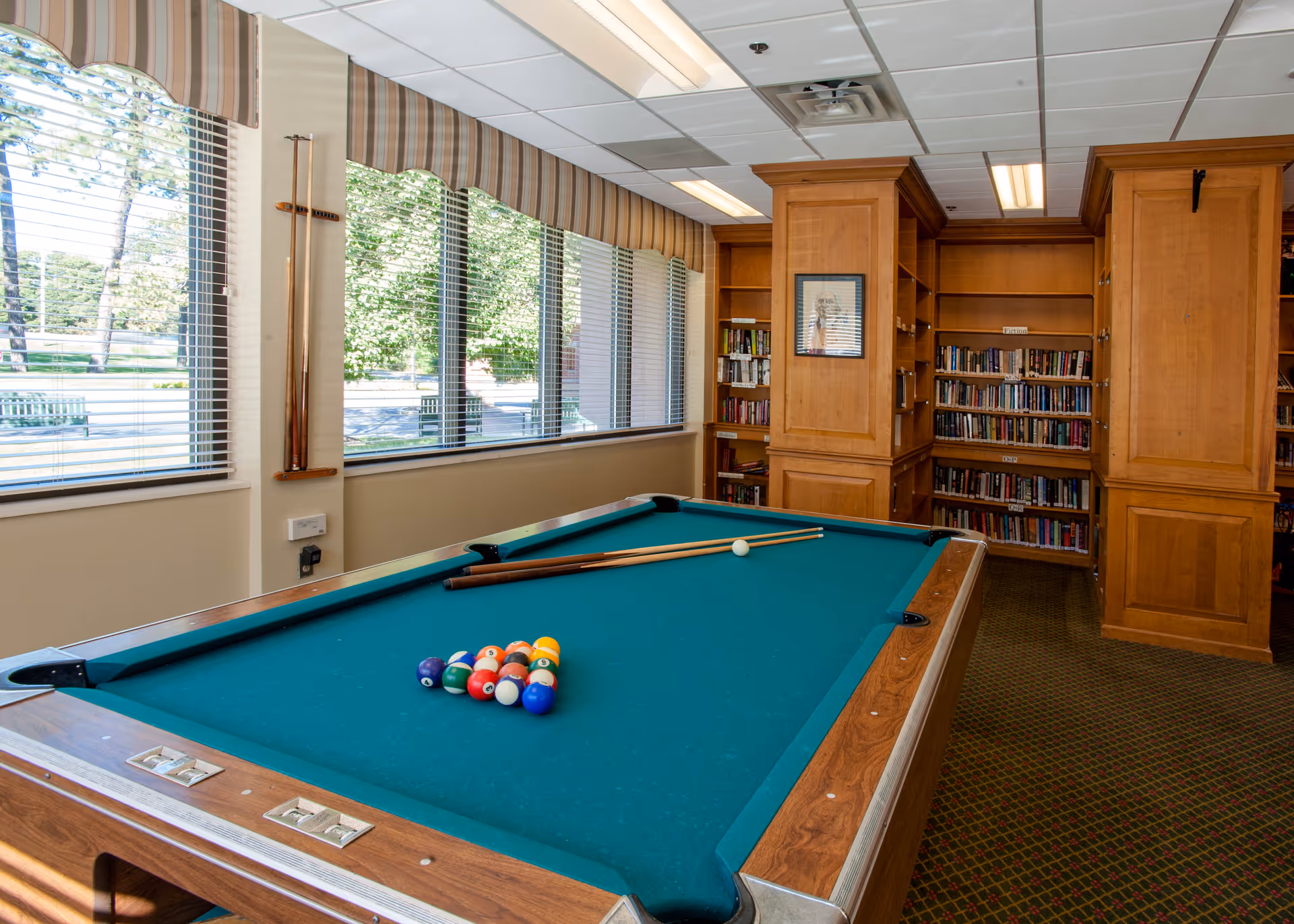 Interior recreation room with a pool table, cues, and bookshelves next to large windows.