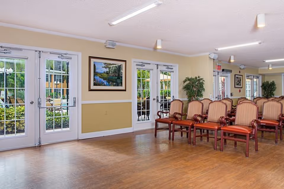 A bright senior living common room with rows of upholstered chairs, wood flooring, and glass doors opening to the outside.