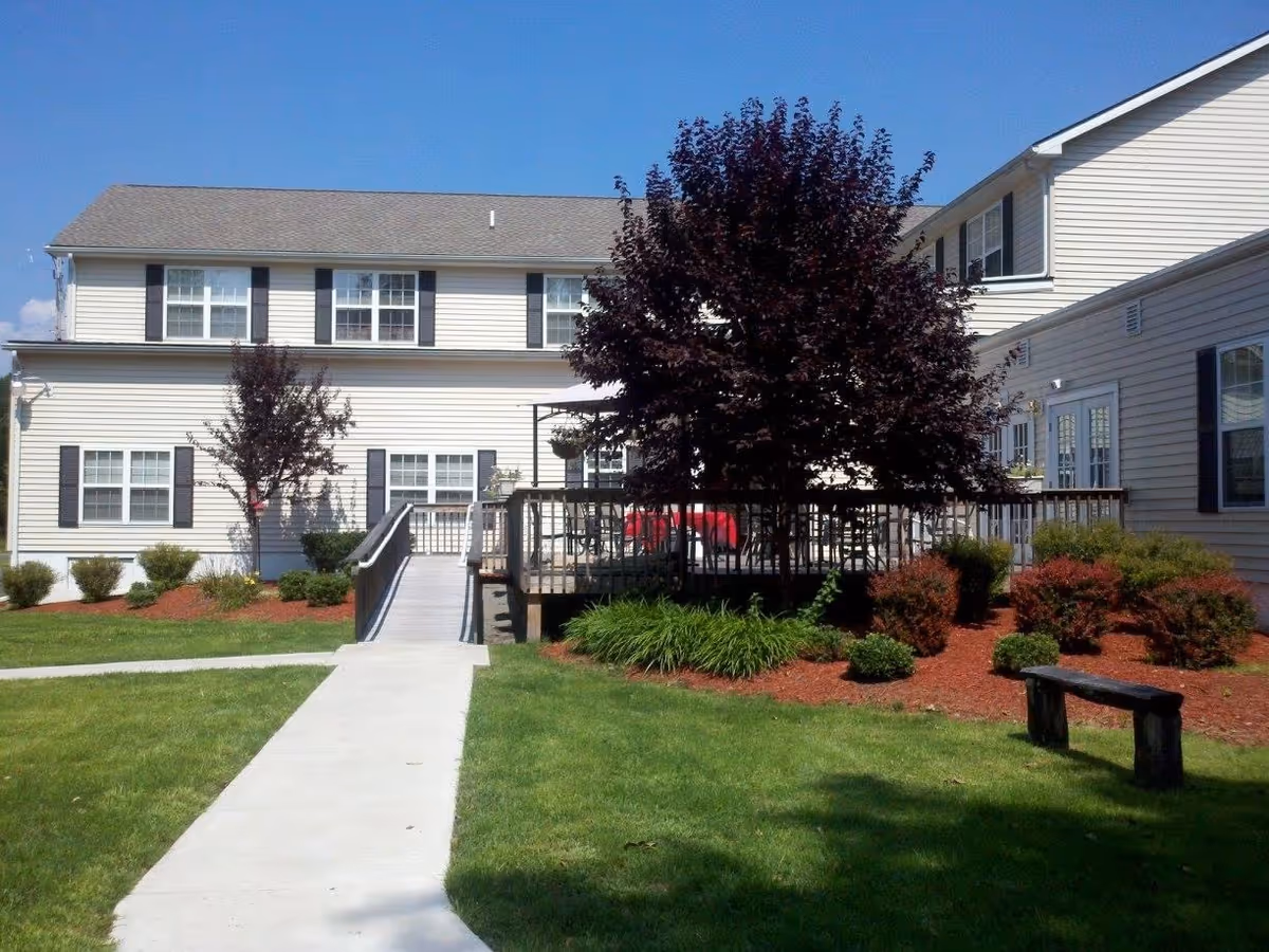 Exterior front of a two-story personal care home with a wheelchair ramp, deck, landscaped lawn and a large tree.