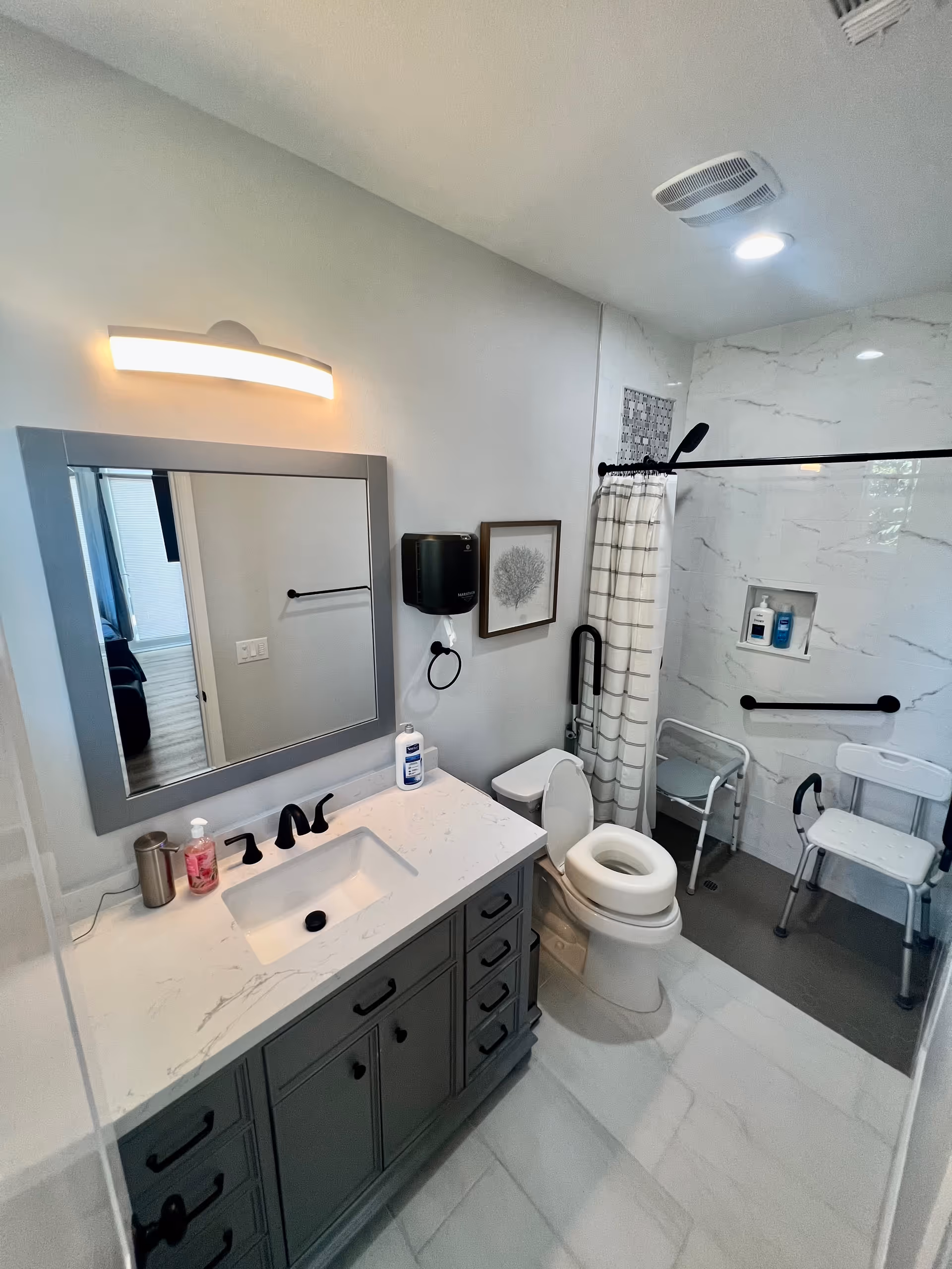 A modern bathroom with a gray vanity featuring a white marble countertop and a rectangular sink. Above the vanity is a large mirror with a light fixture. To the right is a toilet with a raised seat and grab bars for accessibility. Next to the toilet is a walk-in shower with a curtain, a shower chair, and grab bars. The walls of the shower have a marble-like finish and a small built-in shelf holding toiletries.