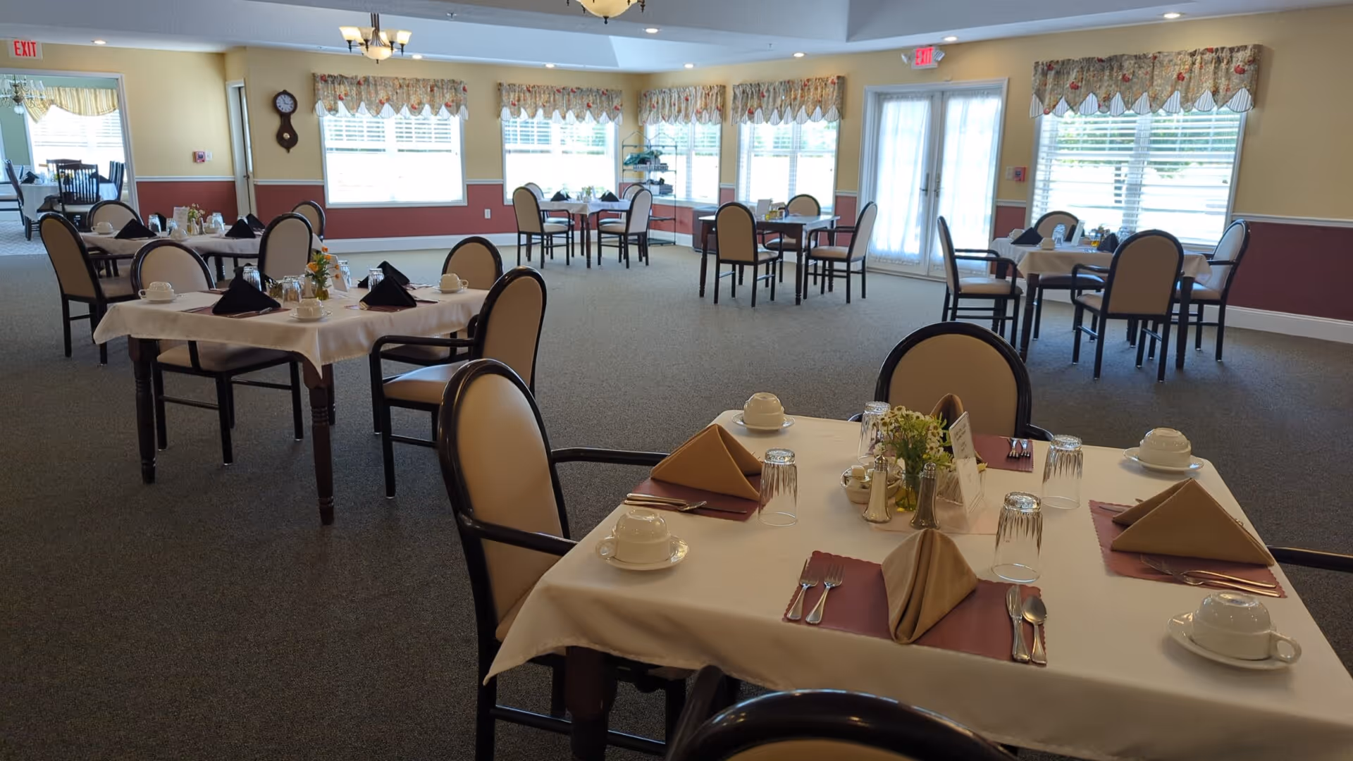 A spacious dining room with multiple tables set for meals, each table covered with a white tablecloth, brown napkins folded in a triangle, cups, glasses, and silverware. The room has large windows with floral valances allowing natural light to fill the space, and chairs with beige cushions surround each table.