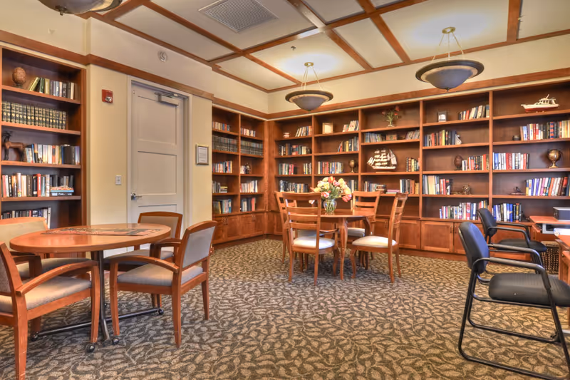 A cozy library or reading room with wooden bookshelves filled with books and decorative items. There are two round wooden tables with chairs around them, a carpeted floor with a leaf pattern, and ceiling lights providing warm illumination.