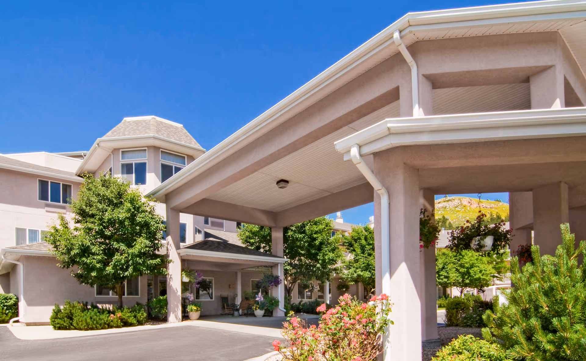 Exterior view of Holiday Hills Estates senior living facility showing a covered entrance with columns, surrounded by green trees and flowering bushes under a clear blue sky.