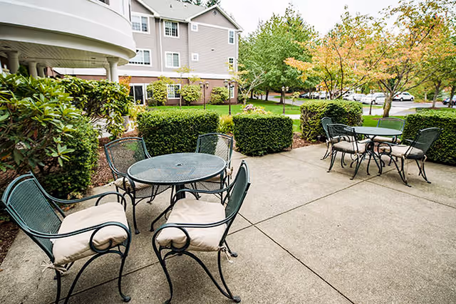Outdoor patio area with metal tables and cushioned chairs on a concrete surface, surrounded by trimmed bushes and trees, with a multi-story residential building in the background.