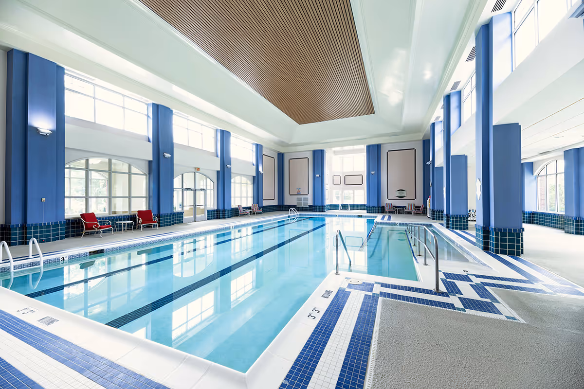 Bright indoor swimming pool with lanes, blue-tiled deck, large windows and seating along the sides.