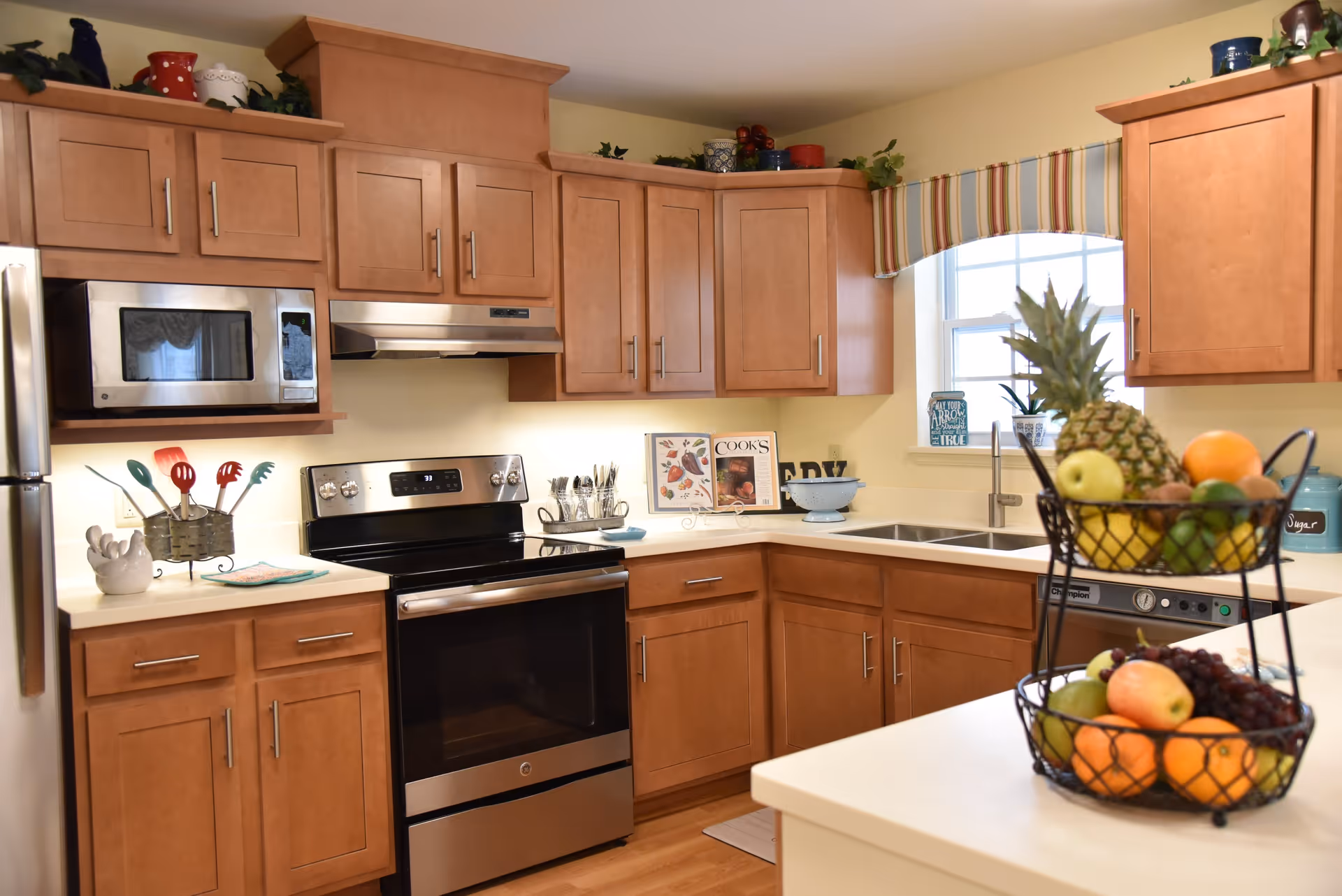 A well-lit kitchen with wooden cabinets, a stainless steel microwave and oven, a double sink under a window with a striped valance, and a two-tiered fruit basket filled with various fruits on the counter.