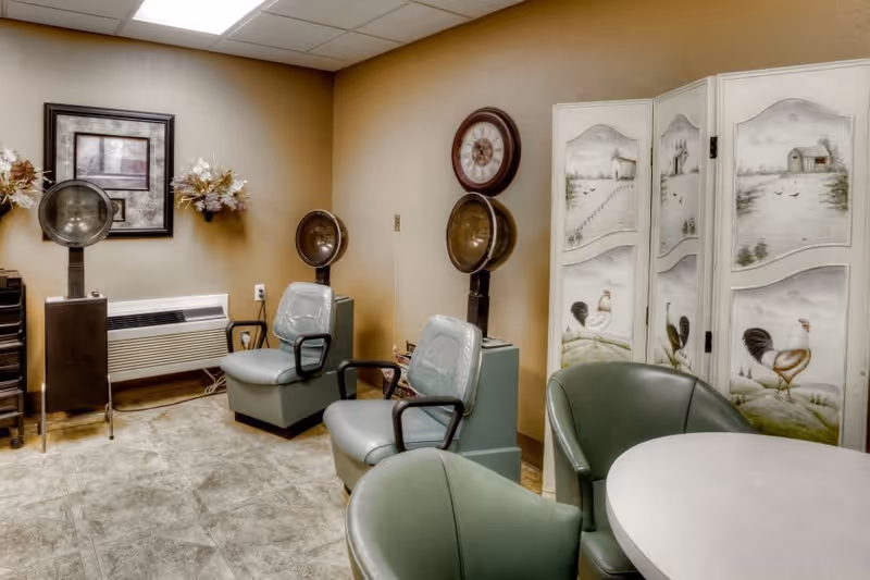 Interior room with two vintage hair dryer chairs, a decorative folding screen with rooster paintings, a round table with green chairs, a wall clock, and framed artwork with floral decorations on the wall.