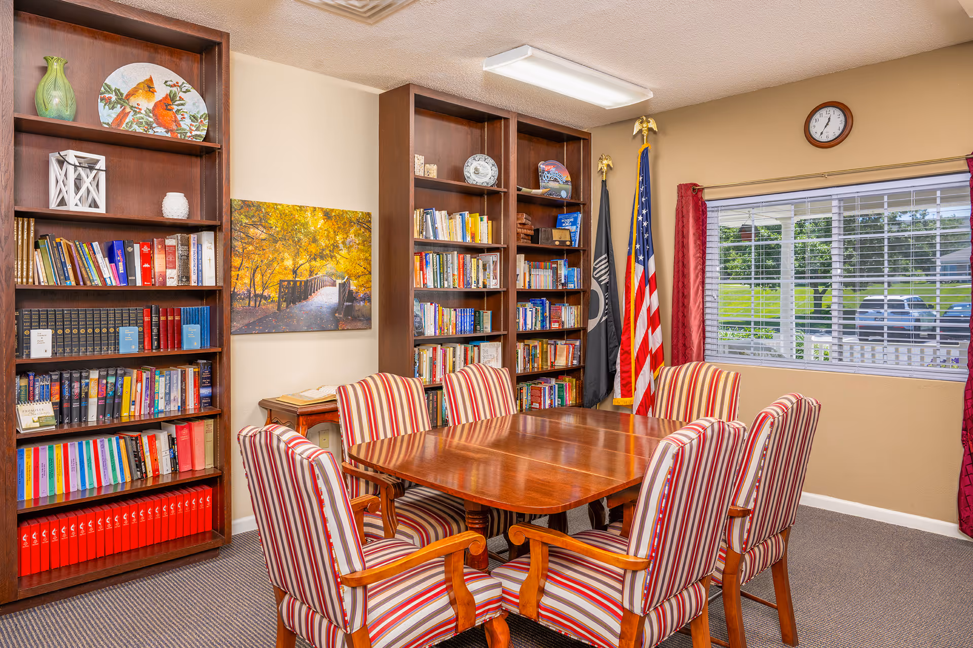 Conference-style room with a wooden table surrounded by striped upholstered chairs, bookshelves filled with books, an American flag, and a window with blinds.