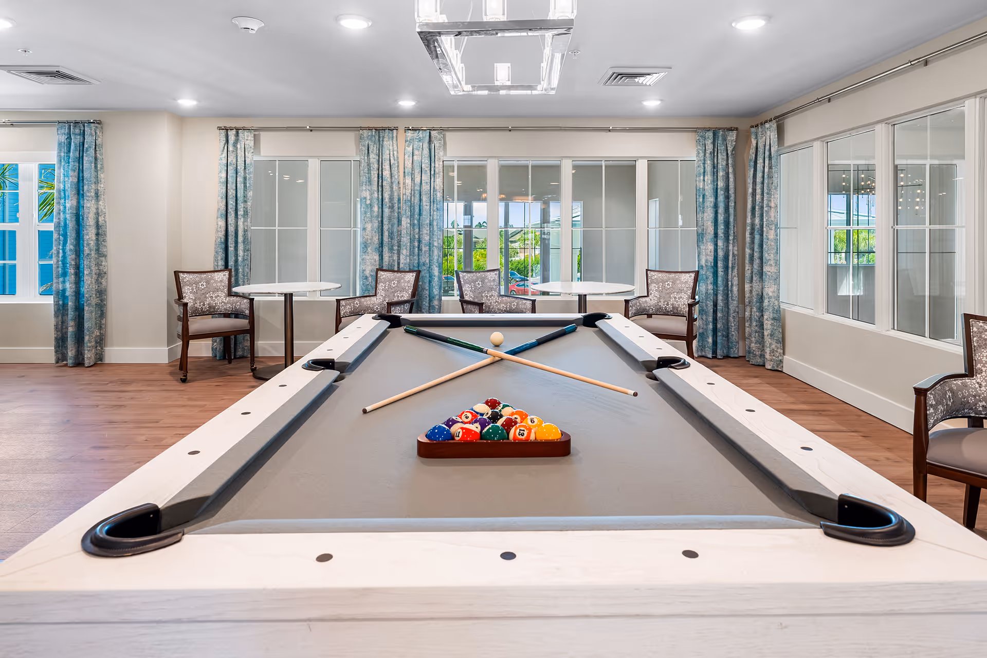 A bright and spacious game room featuring a pool table with billiard balls arranged in a triangle and two crossed cues on the table. The room has wooden flooring, large windows with blue patterned curtains, and several chairs and small round tables along the walls.