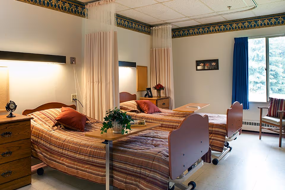 A shared bedroom in a senior living facility with two hospital-style beds, each with striped bedspreads and a red pillow. There are bedside tables with lamps and decorative items, a window with blue curtains, and a chair with a striped cushion. Privacy curtains hang between the beds.