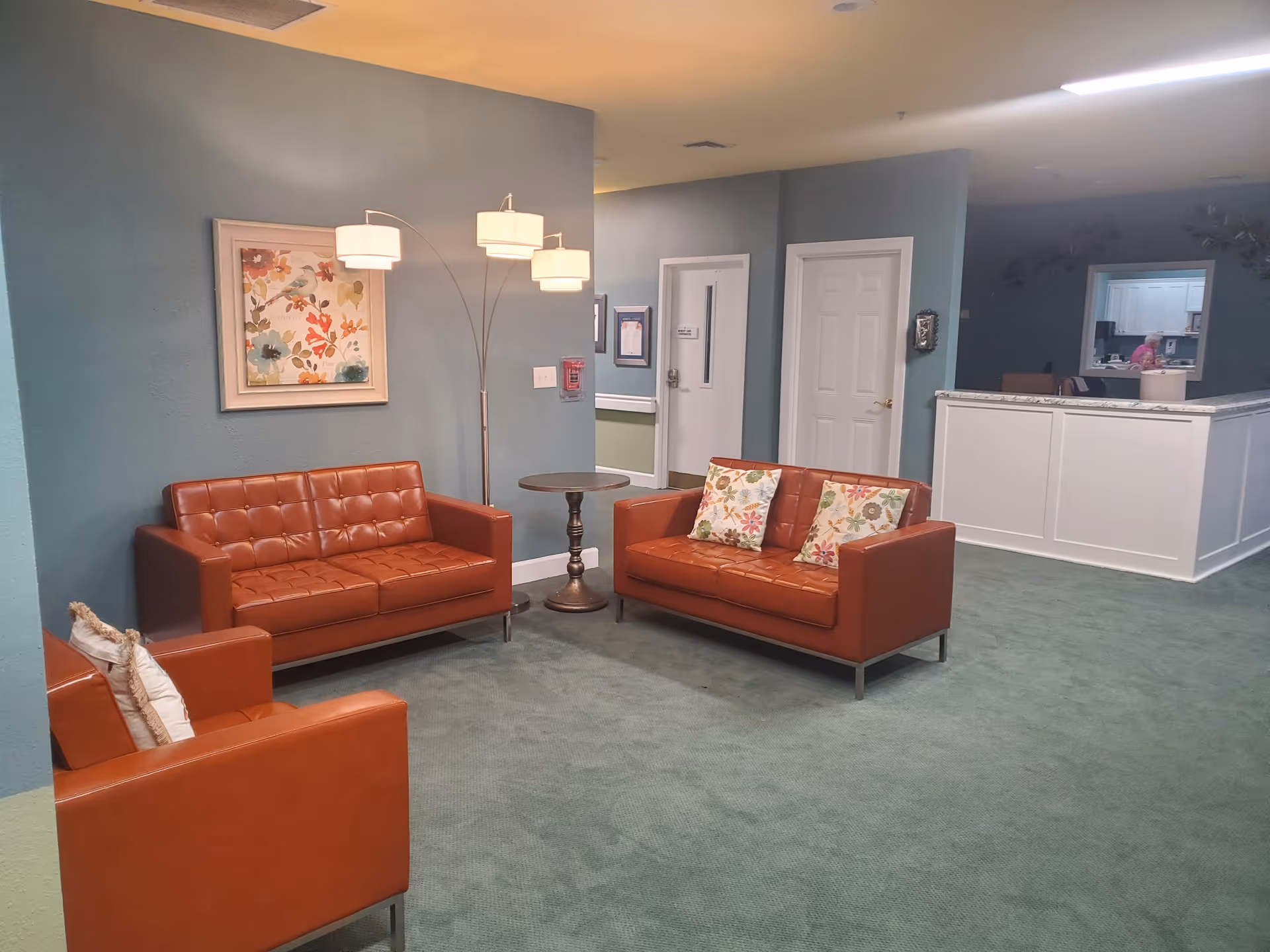 A cozy seating area in a senior living facility with two brown leather sofas and one matching armchair, each with decorative pillows. A floor lamp with four lights stands beside a small round wooden table. The walls are painted blue, and there is a framed floral artwork on the wall. In the background, there is a white counter with a window opening into another room where a person is visible.