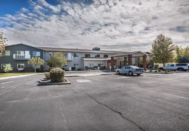 Front exterior of a two-story senior living facility with a covered entrance, parked cars, and a large parking lot under a cloudy sky.