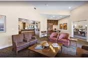 Well-lit assisted living common room with upholstered chairs and sofas around a coffee table, a dining area and bookshelf in the background.