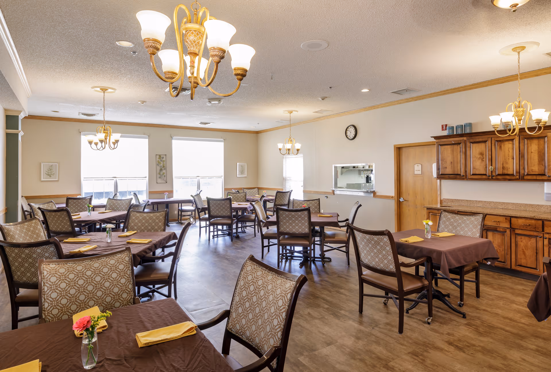 A well-lit dining room in an assisted living facility with multiple tables covered in brown tablecloths, each set with yellow napkins and small flower vases. The room features wooden chairs with patterned upholstery, wooden cabinetry along one wall, and several chandeliers hanging from the ceiling. Large windows allow natural light to fill the space.