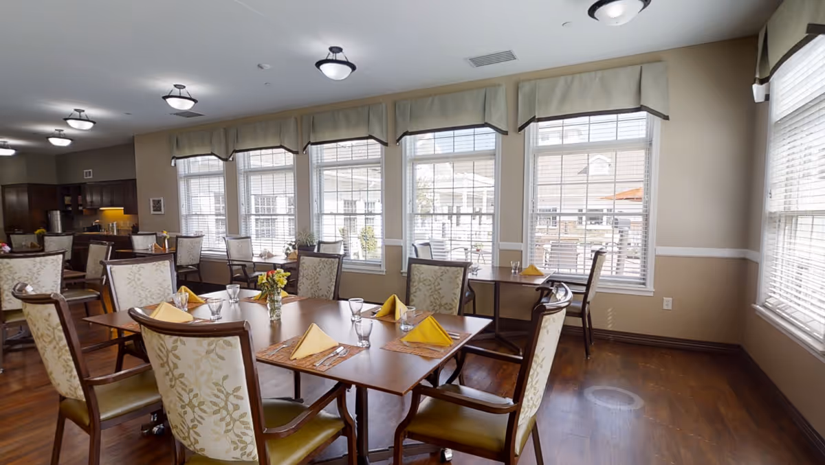 A bright dining room with several wooden tables and cushioned chairs arranged neatly. Each table is set with yellow folded napkins, glasses, and placemats. Large windows with blinds and valances allow natural light to fill the room, and a kitchen area is visible in the background.