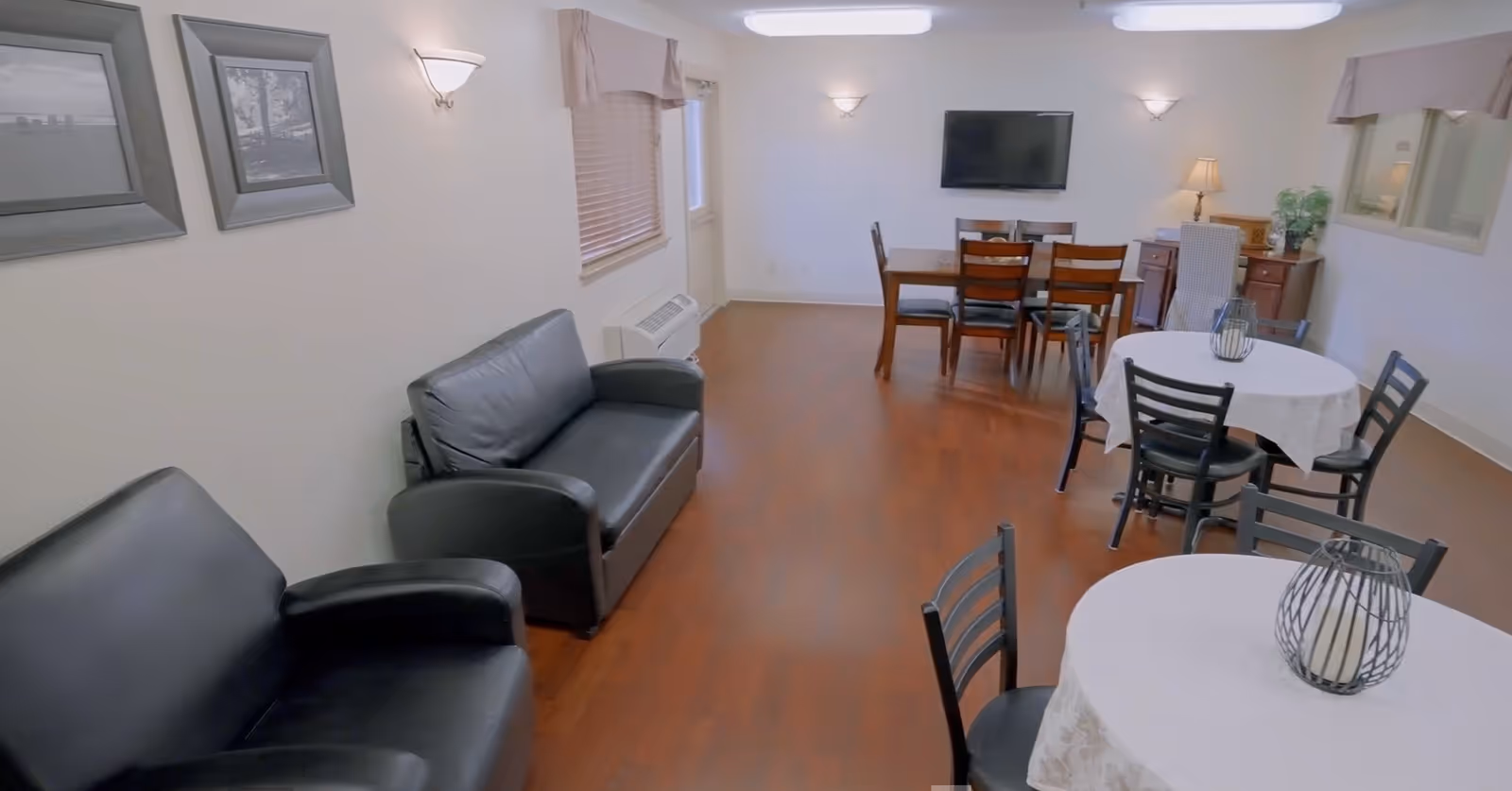 A well-lit common area with wooden flooring featuring two black leather sofas on the left, two round tables with white tablecloths and black chairs, a rectangular dining table with six chairs, a wall-mounted TV, a lamp on a wooden sideboard, and framed pictures on the wall.