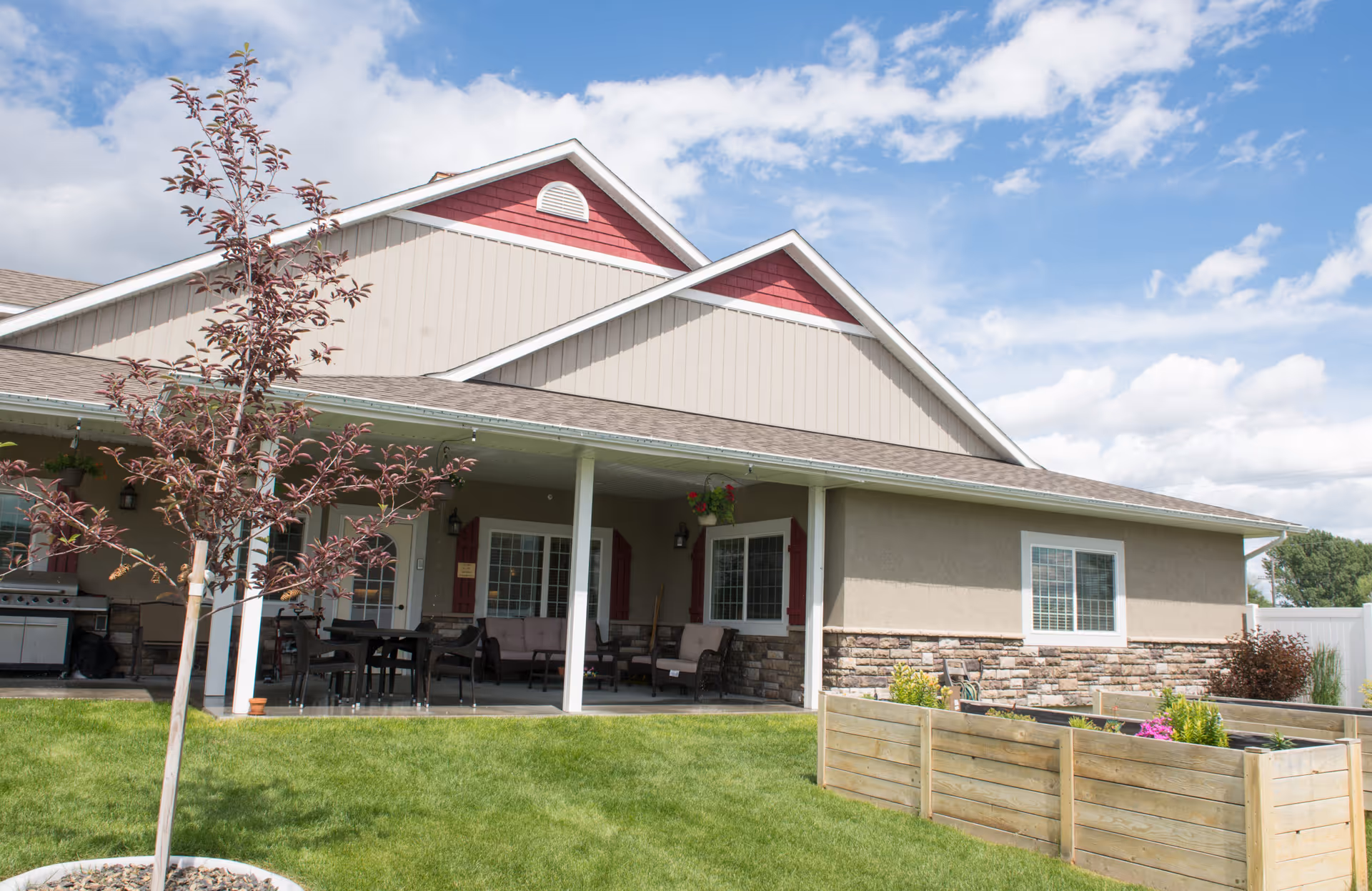 Covered porch and patio of a single-story assisted living building with lawn, raised planter boxes, and outdoor seating under a blue sky.