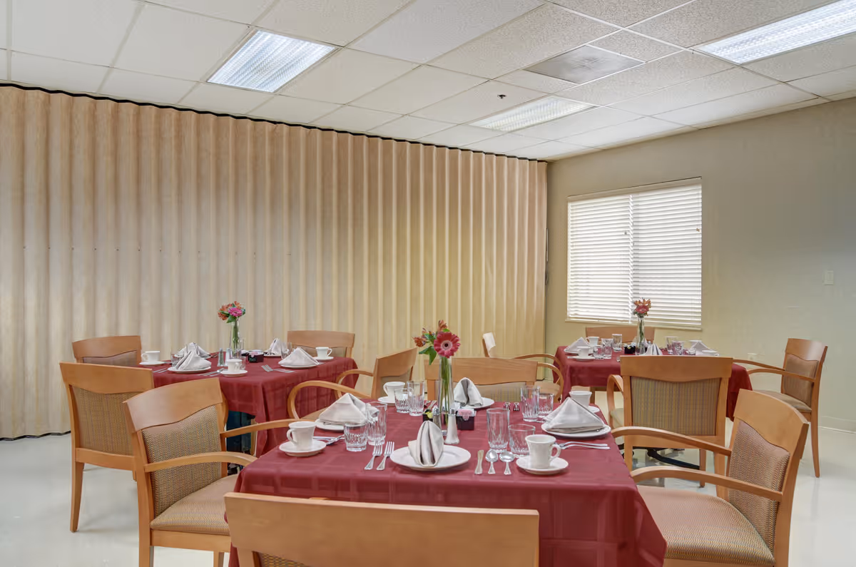 A small dining room with several tables covered in red tablecloths, place settings, and flower centerpieces.