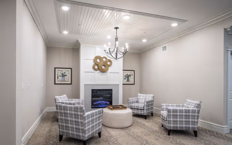 A cozy sitting area with four plaid armchairs arranged around a round beige ottoman in front of a modern electric fireplace. The fireplace is set in a white paneled wall with a decorative circular wall art above it. Two framed floral paintings hang on either side of the fireplace. The ceiling features recessed lighting and a black chandelier with candle-style lights.