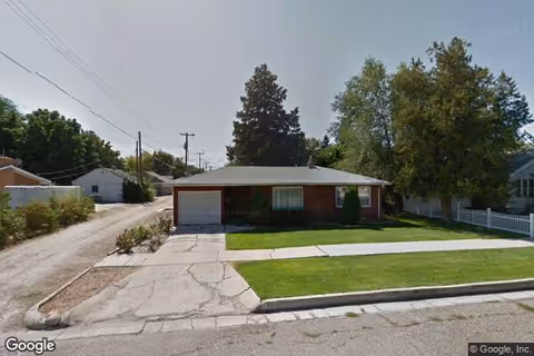 Single-story brick house with a garage on the left side, a driveway, and a well-maintained front lawn with a sidewalk. Trees and neighboring houses are visible in the background under a clear sky.