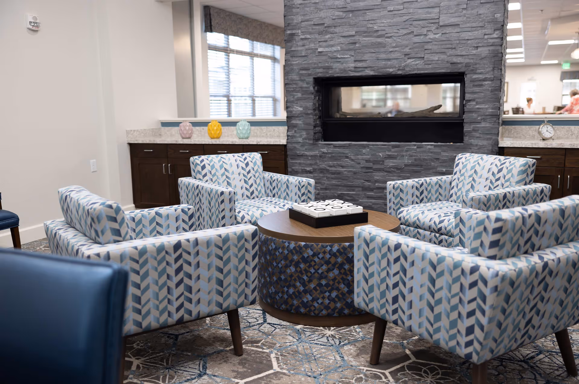 A cozy seating area with four patterned armchairs arranged around a round wooden table with a tic-tac-toe game on top. Behind the seating area is a modern stone fireplace and a countertop with decorative vases and a clock. Large windows with blinds allow natural light into the room.