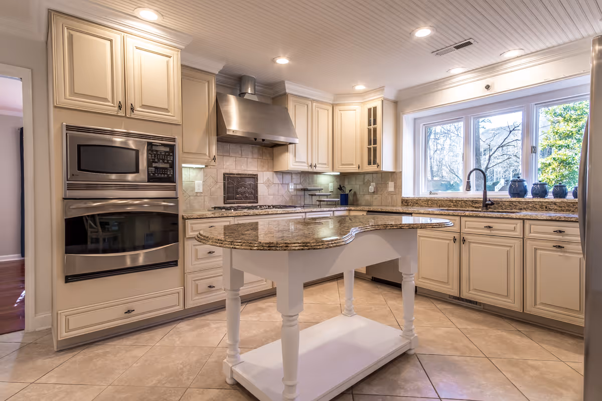 Bright and spacious kitchen with cream-colored cabinets, stainless steel oven and microwave, a large window over the sink letting in natural light, and a central island with a granite countertop.