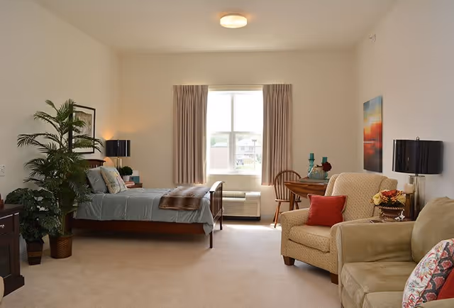 A well-lit senior living bedroom featuring a single bed with light blue bedding and a brown throw blanket, a wooden nightstand with a lamp and books, a large potted plant, a window with beige curtains, a small round wooden table with two chairs, an armchair with a red pillow, a beige sofa with a floral pillow, and a colorful painting on the wall.