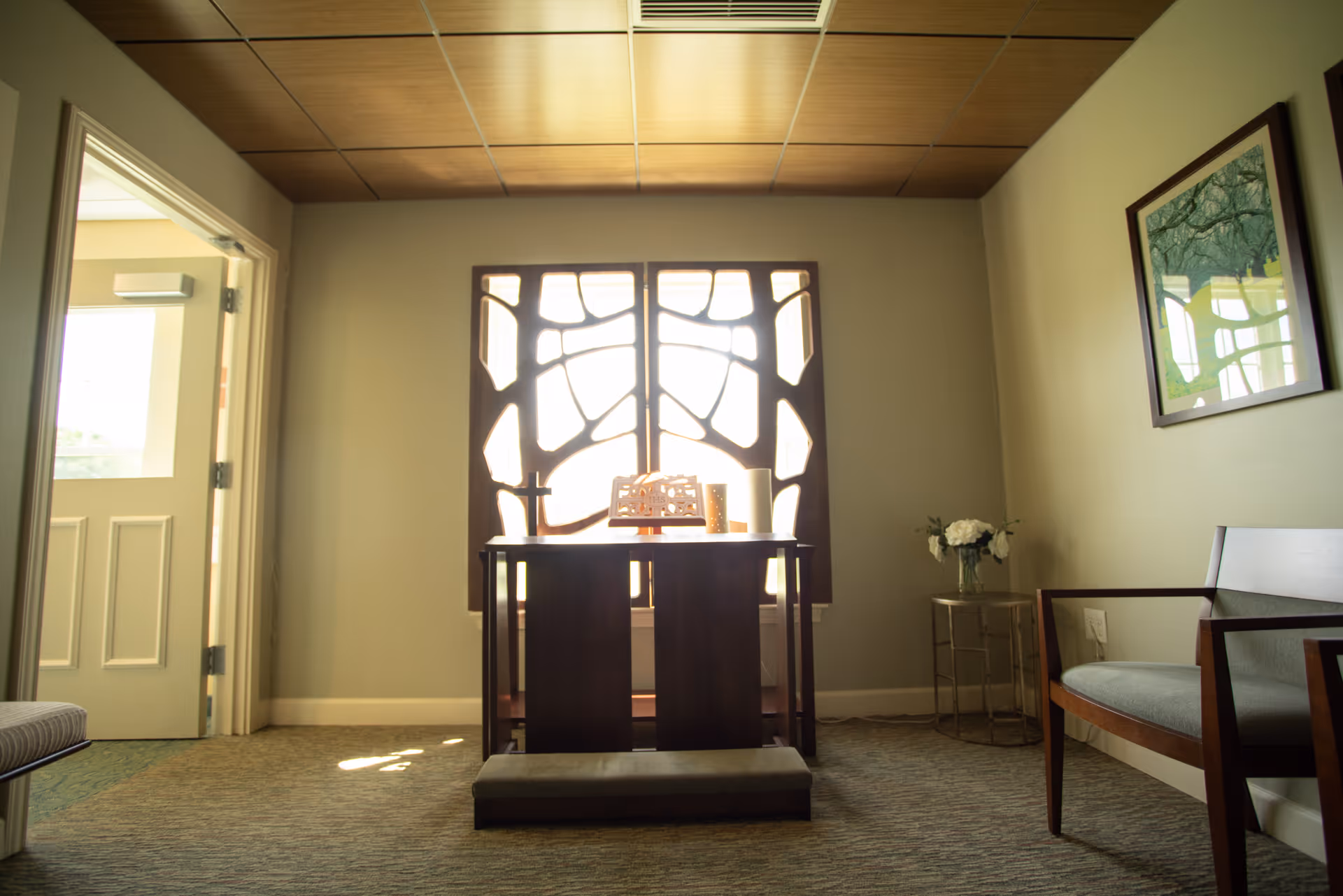 A quiet room with a decorative wooden table in front of a window with artistic wooden panels. The room has a wooden ceiling, a small cushioned bench in front of the table, a side table with a vase of white flowers, and a wooden chair with a cushion. A framed picture hangs on the right wall, and an open door is visible on the left side.