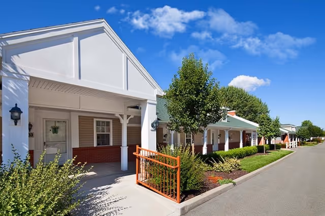 Exterior view of Longwood At Oakmont Retirement Community showing a row of single-story buildings with covered entrances, green roofs, and well-maintained landscaping under a blue sky with some clouds.