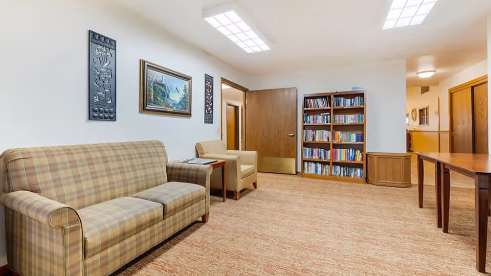 A cozy common area with a plaid sofa, beige armchair, wooden side table, and a bookshelf filled with books. The walls are decorated with framed artwork and decorative panels. The room has a carpeted floor and bright overhead lighting.