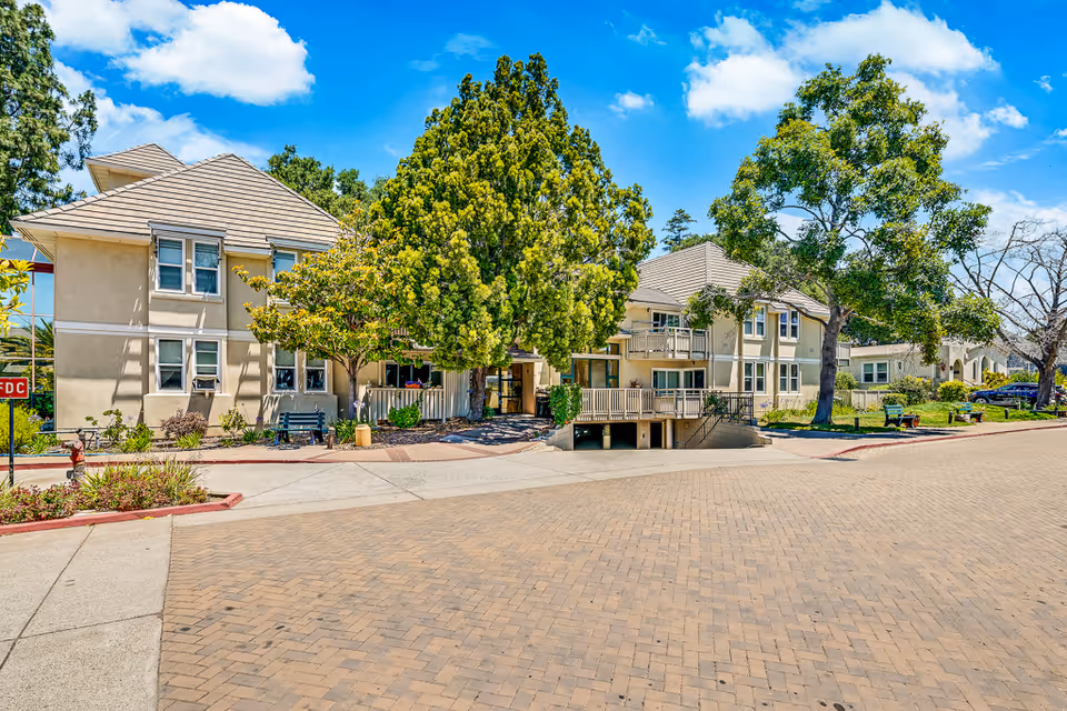 Exterior view of a senior living facility with beige buildings, multiple windows, and a tiled roof. The scene includes a paved driveway, green trees, benches, and a clear blue sky with some clouds.