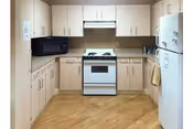 A clean kitchen with light wood cabinets, a white stove with oven, a white refrigerator, a black microwave, and light wood flooring.