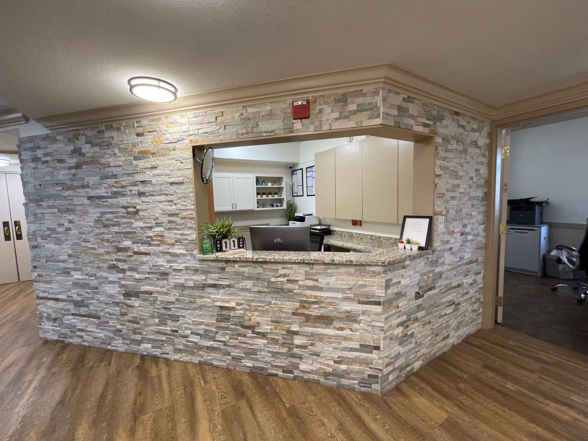 Reception area with a stone accent wall and a granite countertop window opening into an office space. The office has beige cabinets, a computer, and some small plants and decor on the counter. The floor is wood, and there is a door leading to another room with office equipment visible.