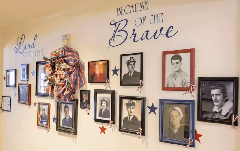 A wall display featuring framed black and white and color photographs of military veterans, decorated with red, white, and blue stars and ribbons. The wall has the phrases 'Land of the Free' and 'Because of the Brave' written in large decorative letters, along with a patriotic wreath.