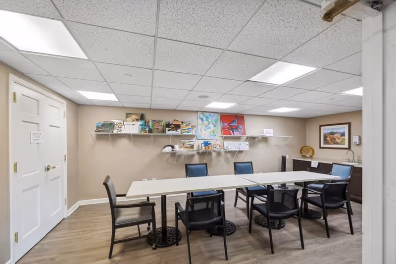 A small meeting or activity room with a rectangular table surrounded by six chairs. The walls are beige with a white ceiling featuring recessed lighting. There are two shelves mounted on the wall holding various books, artwork, and papers. A framed picture hangs on the right wall above a counter with a sink and a soap dispenser. The floor is wood laminate, and there is a closed white double door on the left side of the room.