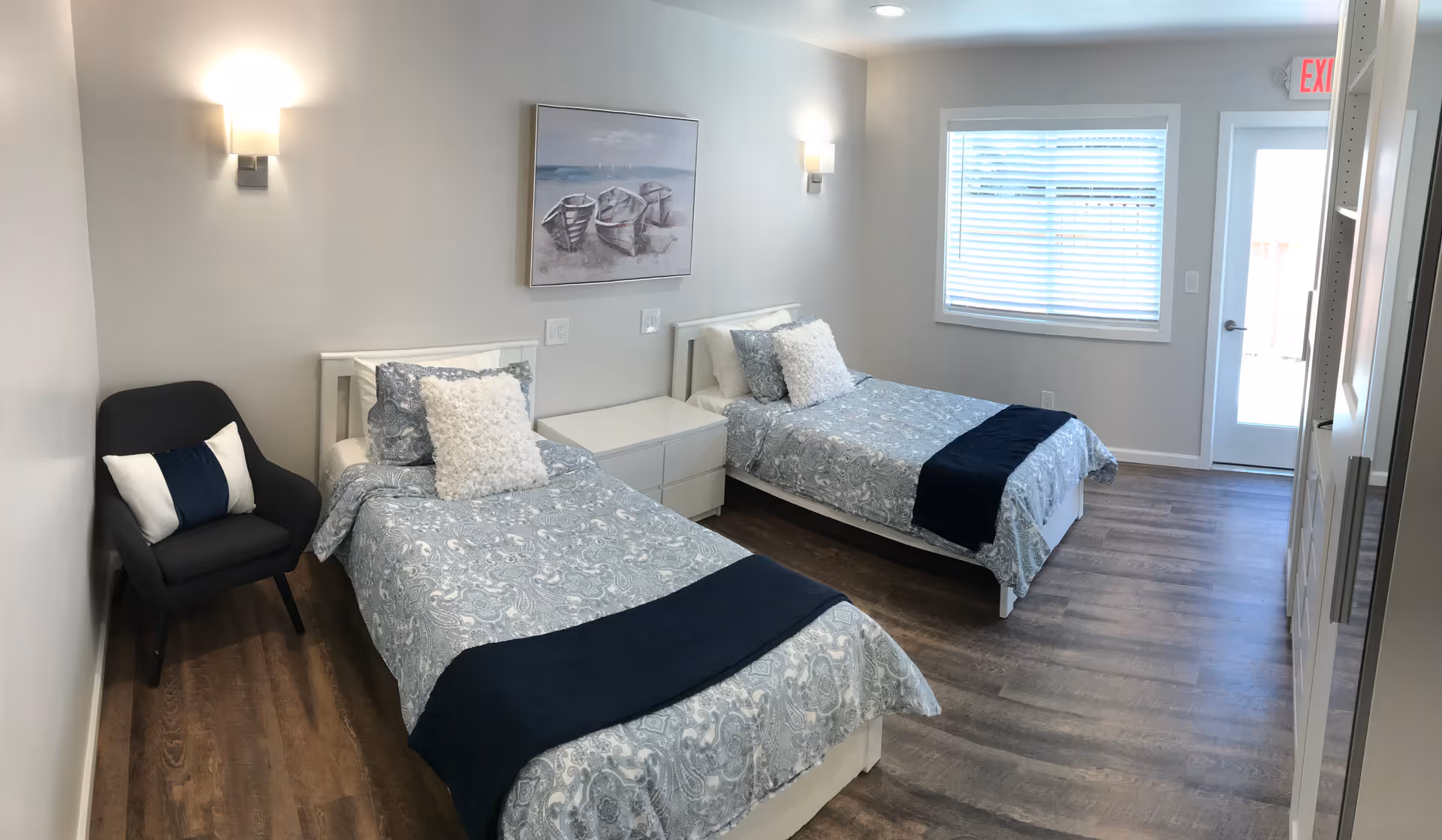 A bright bedroom with two single beds, each with patterned blue and white bedding and a dark blue throw blanket at the foot. Between the beds is a white nightstand. A dark gray armchair with two pillows is positioned in the corner. The walls are light gray with two wall-mounted lights and a painting of three boats above the nightstand. A window with blinds and a door with an exit sign above it are visible on the far wall. The floor is wood with a medium brown finish.