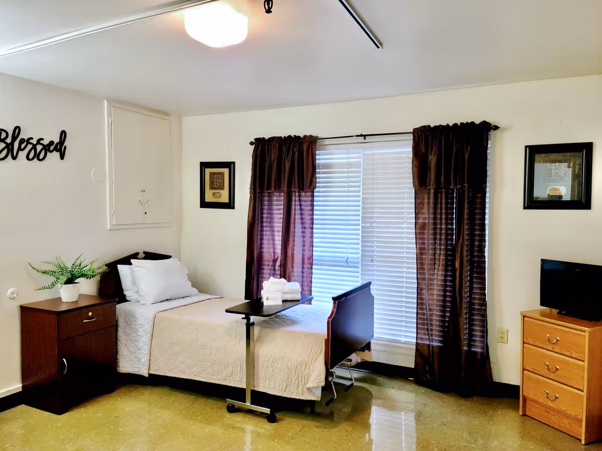 A simple, clean bedroom in a nursing facility with a single bed covered in a beige quilt, a small bedside table with a potted plant, a rolling overbed table with folded towels, dark brown curtains covering a window with blinds, a small TV on a wooden dresser, and framed artwork on the walls. The word 'Blessed' is displayed on the wall above the bedside table.