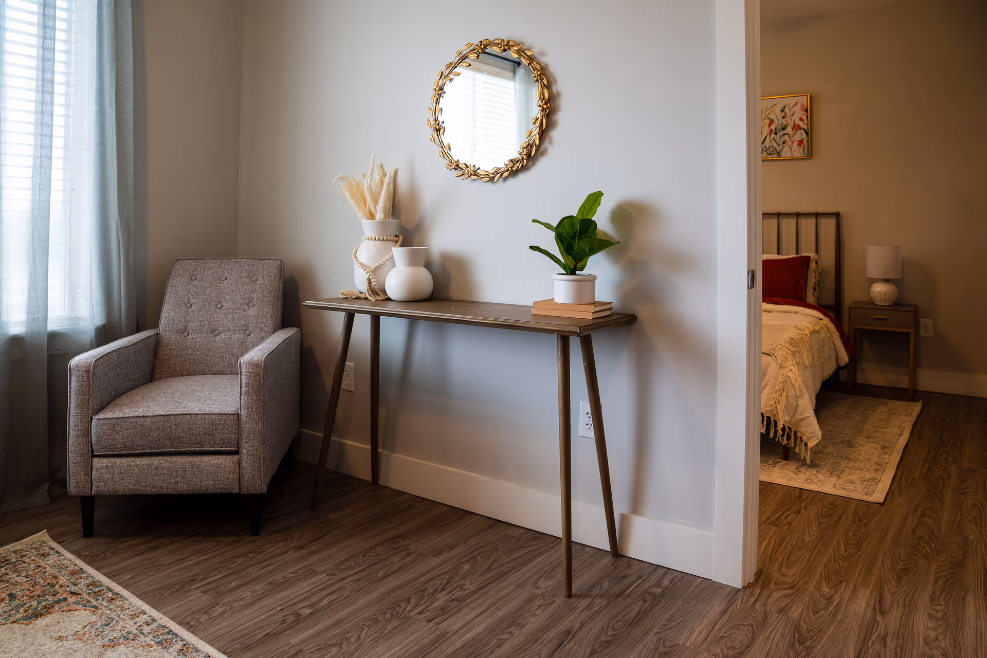 A cozy corner of a living area featuring a gray upholstered armchair next to a window with sheer curtains. A narrow wooden console table against the wall holds decorative items including a white vase with dried pampas grass, another white vase, and a small potted plant on top of two books. Above the table hangs a round mirror with a decorative gold frame. To the right, an open doorway reveals part of a bedroom with a bed, nightstand, lamp, and wall art.