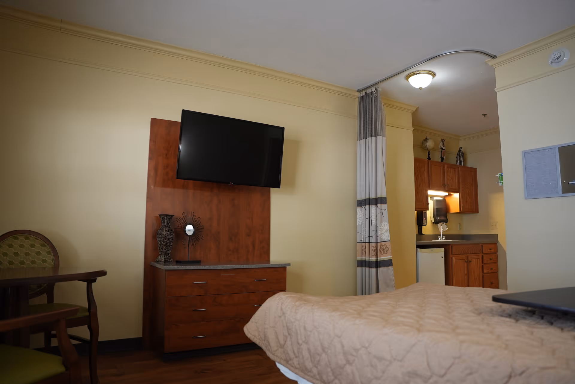 Studio-style bedroom in a senior living facility with a bed in the foreground, wall-mounted TV above a wooden dresser, a small table and chair, and a kitchenette area behind a curtain.