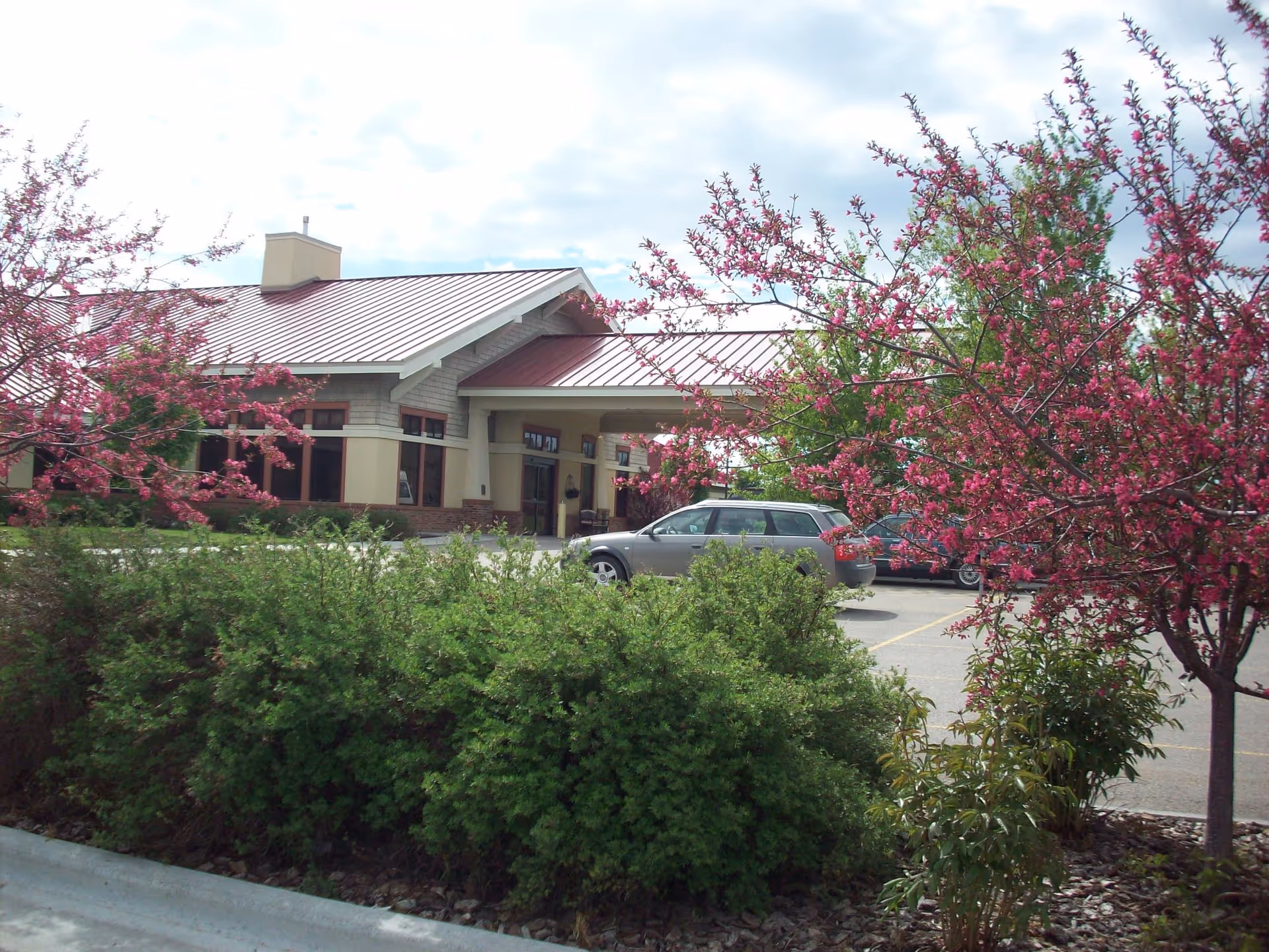 Front entrance of a senior living facility with a covered drop-off, parked cars, and blooming pink trees in the foreground.